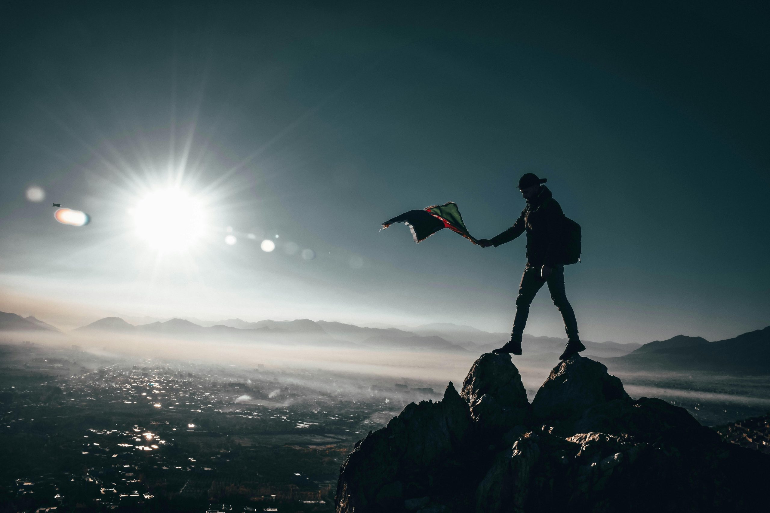 A climber holds a flag on a rocky peak, silhouetted against a misty mountain landscape in the morning sun.